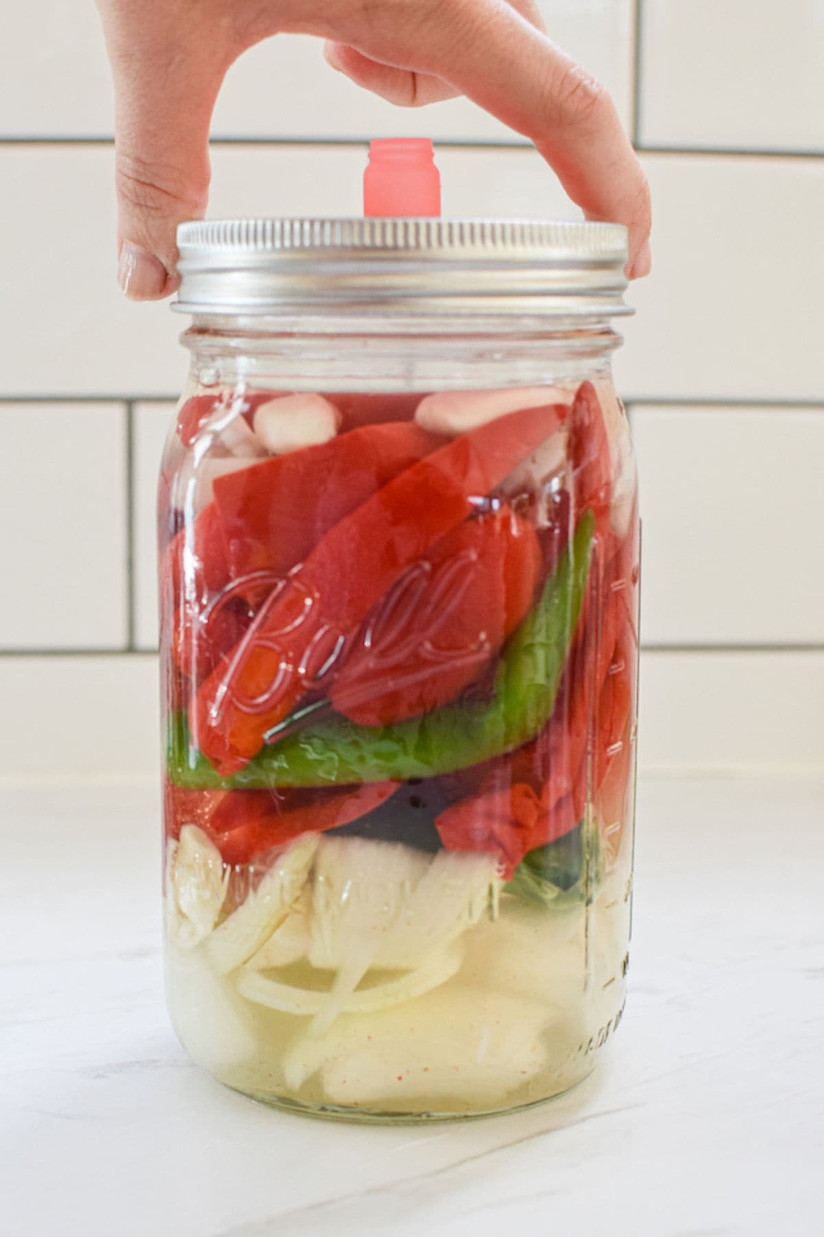 A hand holds a large glass jar of fermented hot sauce ingredients—sliced onions, red and green peppers, and garlic cloves—in brine, sealed with a metal lid and pink airlock, set against a white tiled background.