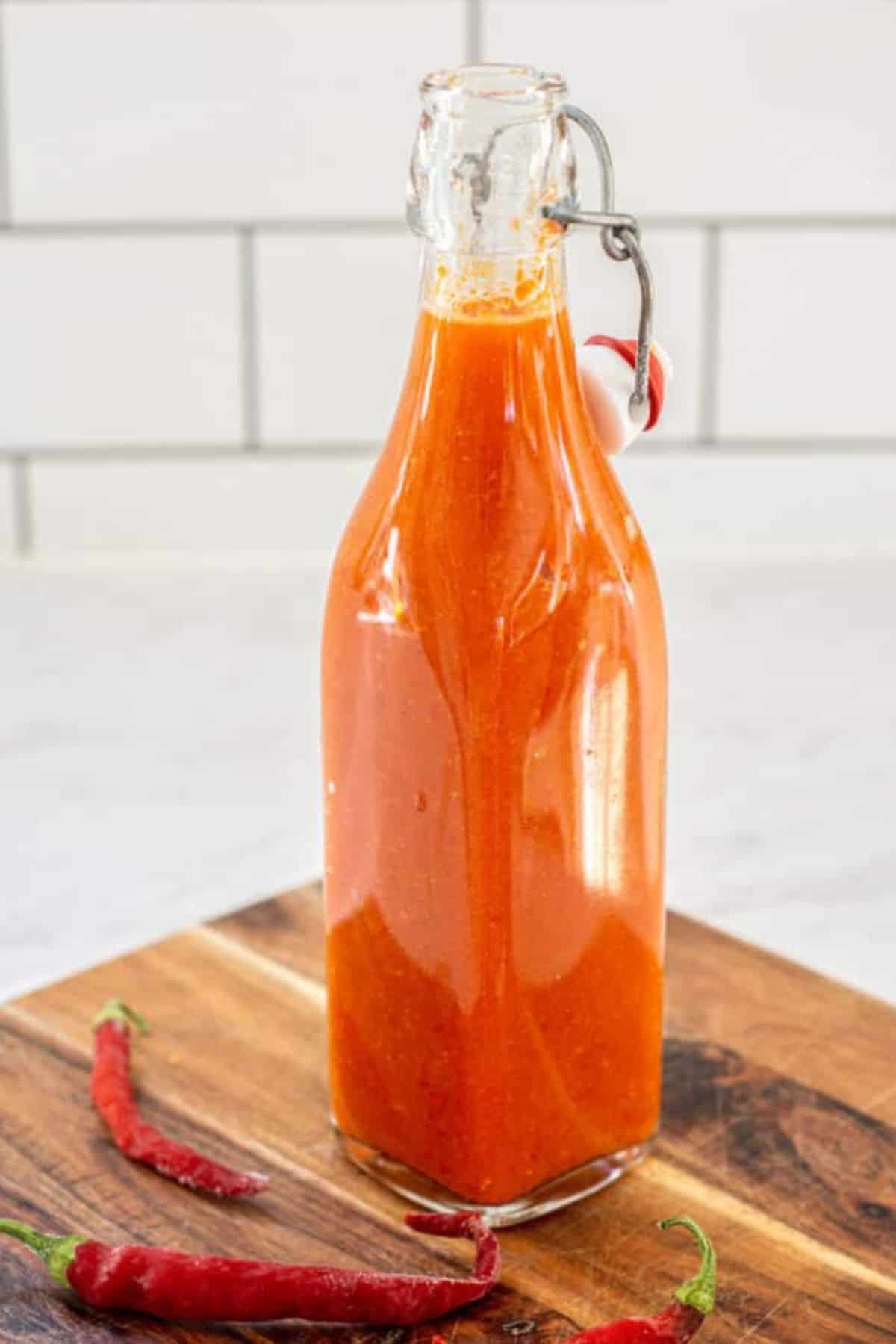 A glass bottle filled with bright orange-red fermented hot sauce sits on a wooden board, with two red chili peppers lying beside it. The background is a white tiled wall.