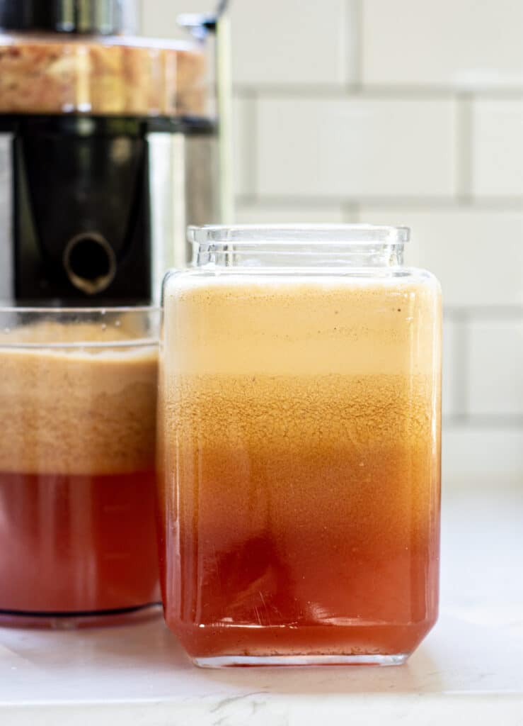 two jugs of apple juice, with apple foam at the top and a juicer in the background