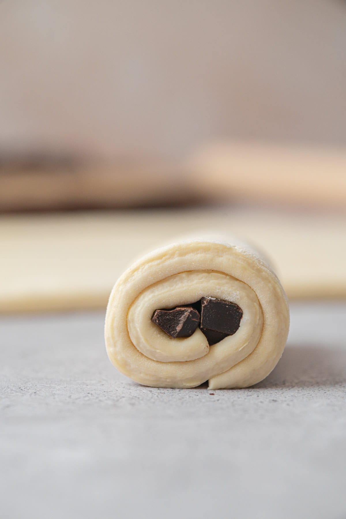 A close-up of an unbaked sourdough pain au chocolat roll with visible layers of dough and chunks of dark chocolate in the center, resting on a light gray surface.
