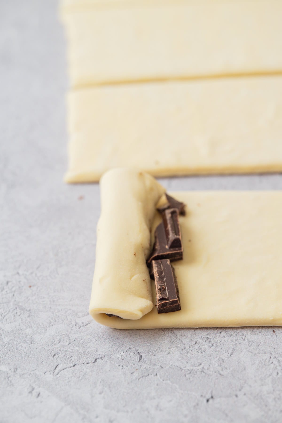 A sheet of sourdough pain au chocolat dough is being rolled up with pieces of chocolate inside on a light gray surface, with more dough strips visible in the background.