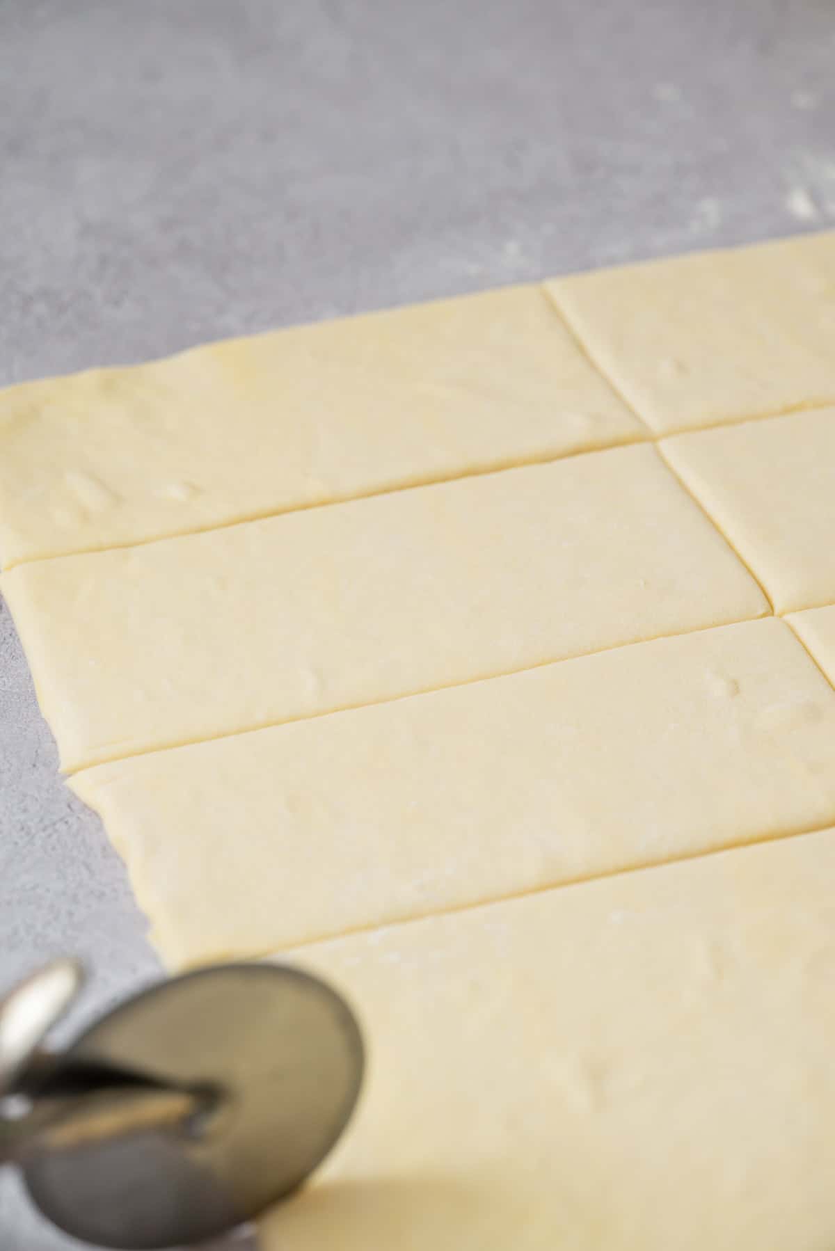 A sheet of sourdough pain au chocolat dough on a gray surface is being cut into rectangles with a metal pizza cutter.