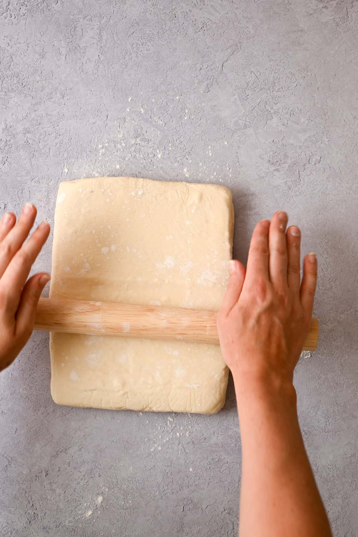 Two hands use a wooden rolling pin to roll out a rectangular piece of sourdough pain au chocolat dough on a light gray, lightly floured surface.
