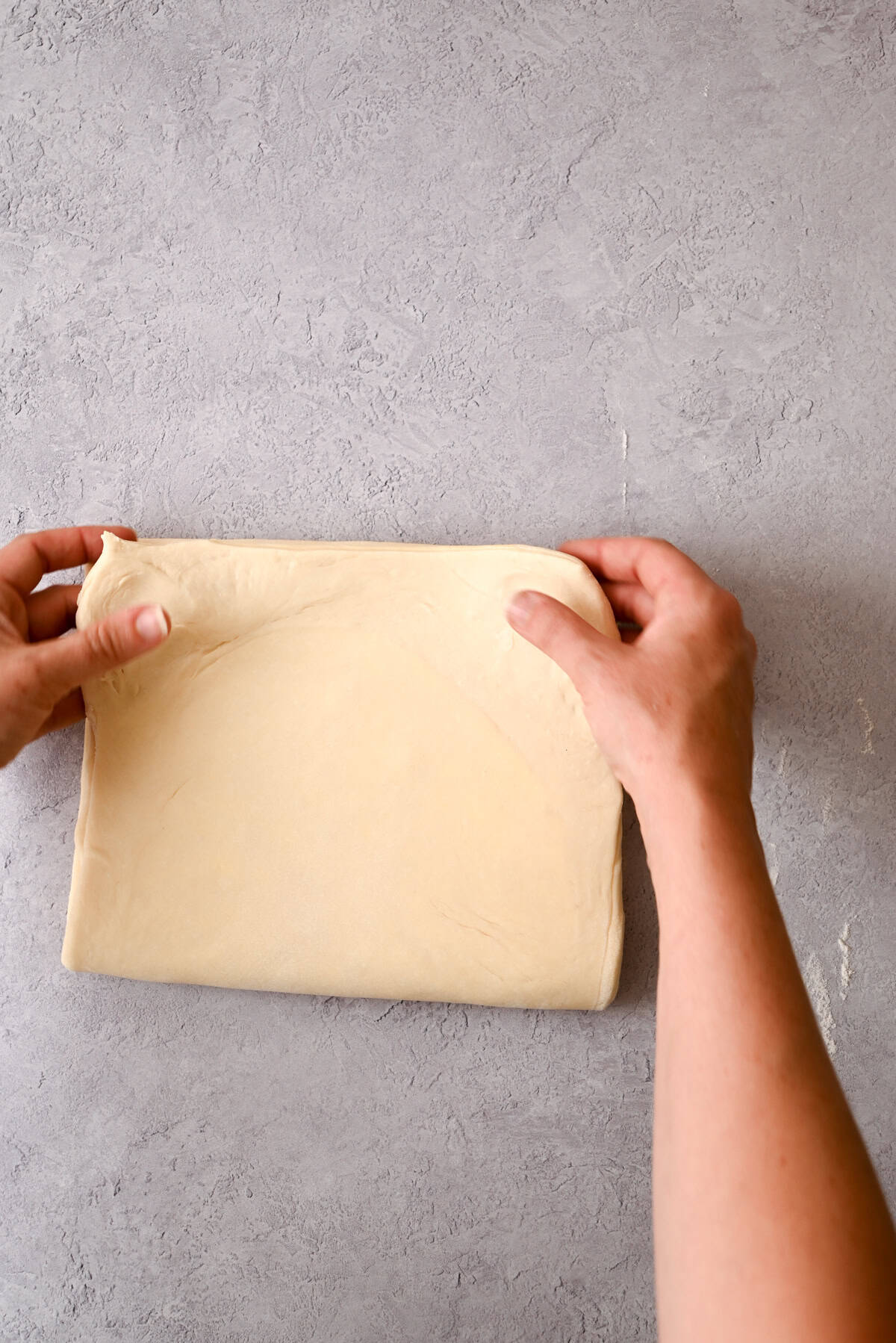 Two hands are folding a sheet of uncooked sourdough pain au chocolat dough on a light grey countertop.