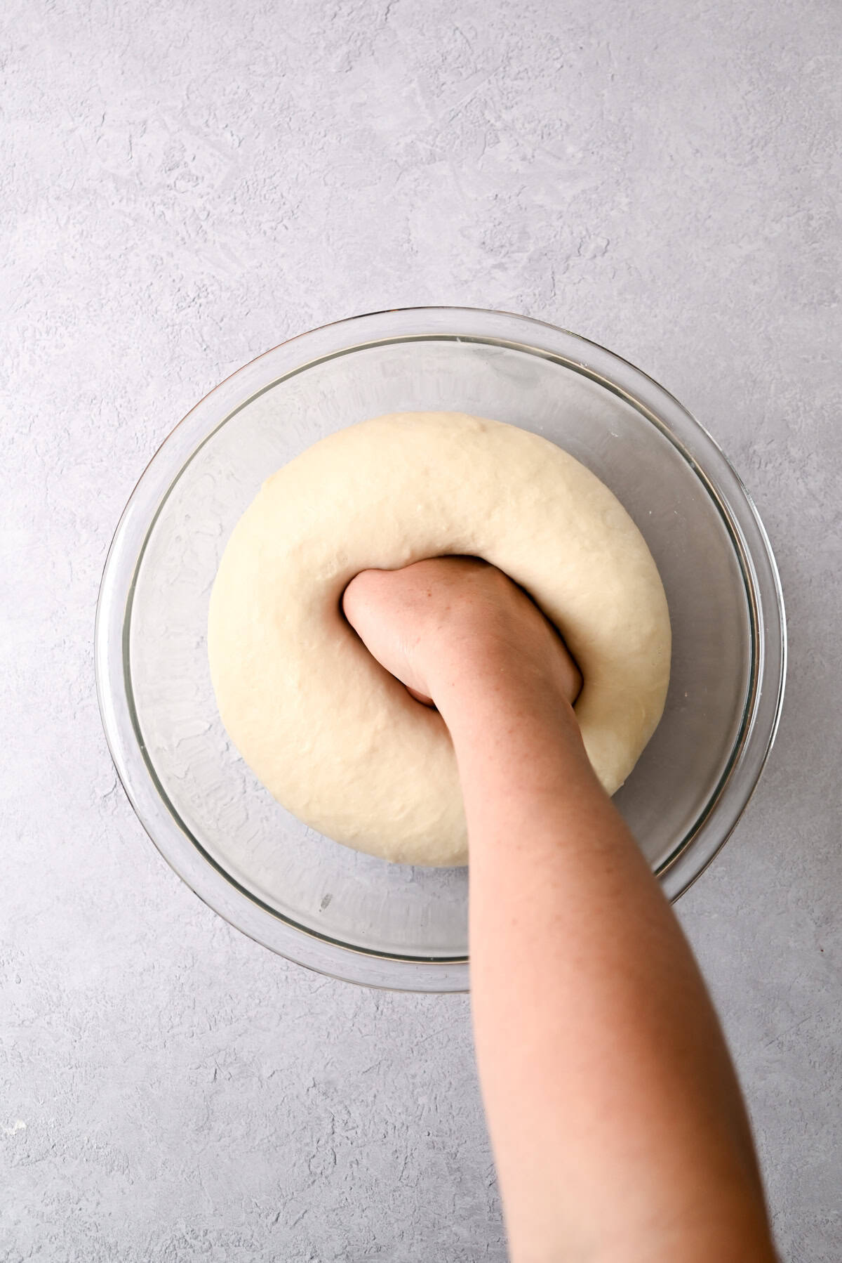 A hand is punching down risen sourdough pain au chocolat dough in a glass bowl on a light gray surface, showing the process of making bread.