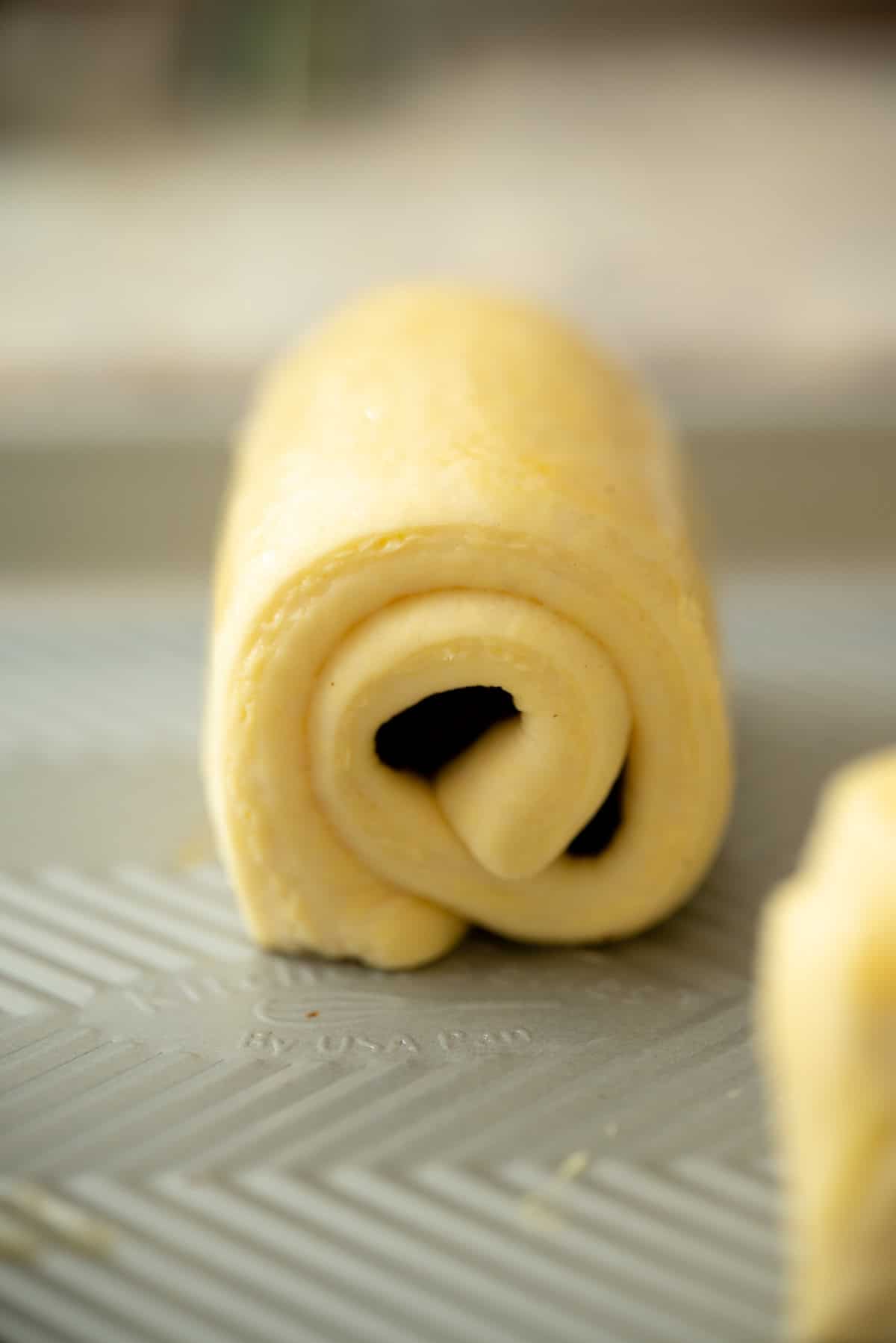 A close-up of an unbaked, rolled sourdough pain au chocolat pastry dough on a textured baking sheet, with a glossy, egg-washed surface. The spiral layers of the dough are clearly visible.