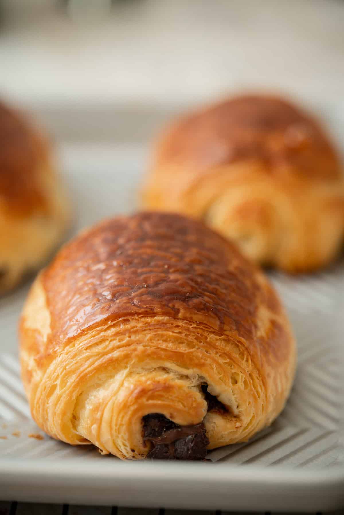 A close-up of a golden, flaky sourdough pain au chocolat pastry on a patterned plate, with two more pastries blurred in the background.