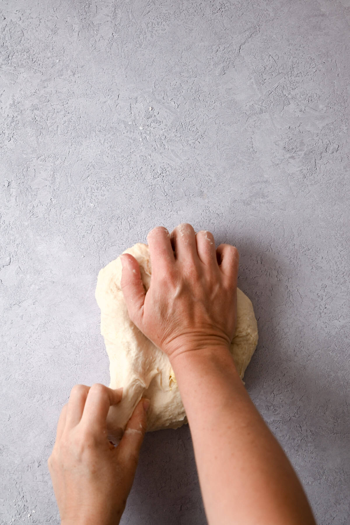 Two hands are kneading a ball of sourdough pain au chocolat dough on a light grey, textured surface. The dough appears soft and slightly sticky, and the process of kneading is in progress.