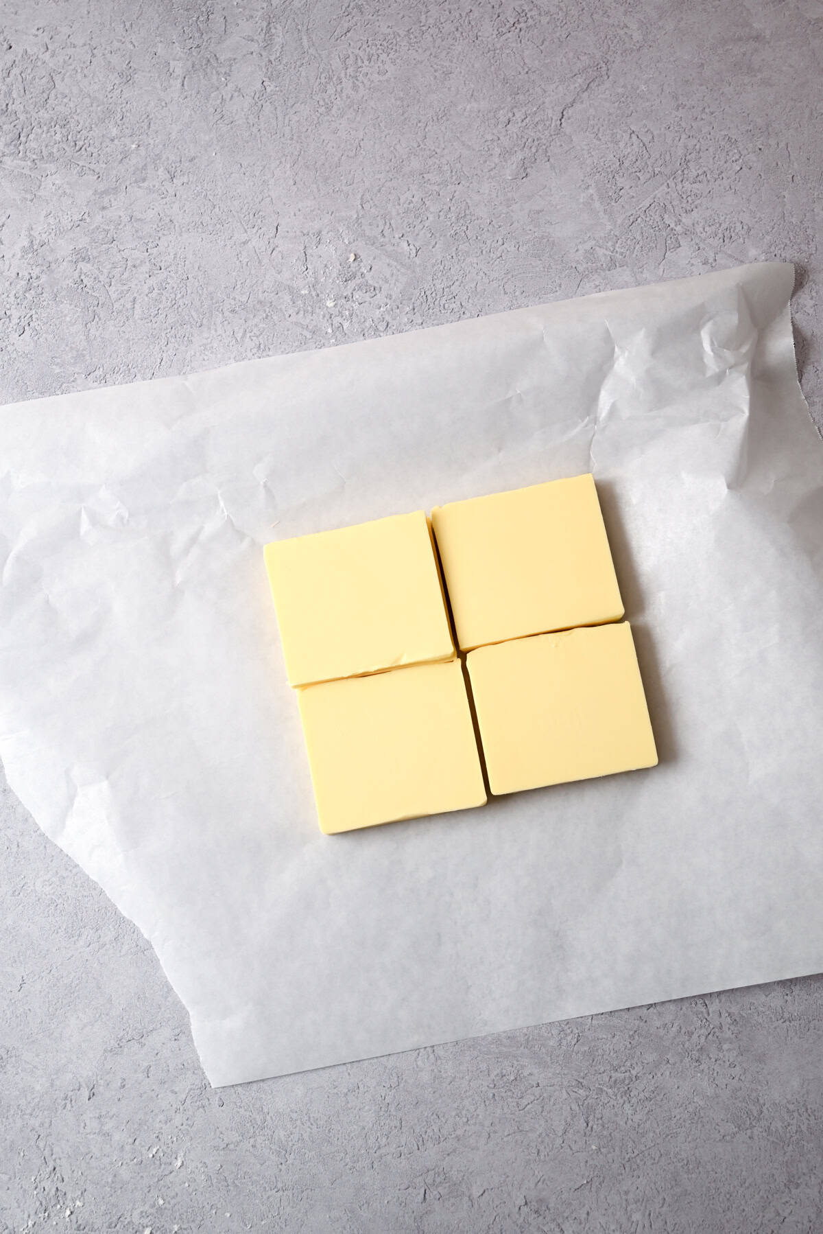 Four rectangular pieces of butter are arranged in a square on an unwrapped sheet of white parchment paper, set on a light grey textured surface—perfect for creating homemade sourdough pain au chocolat.