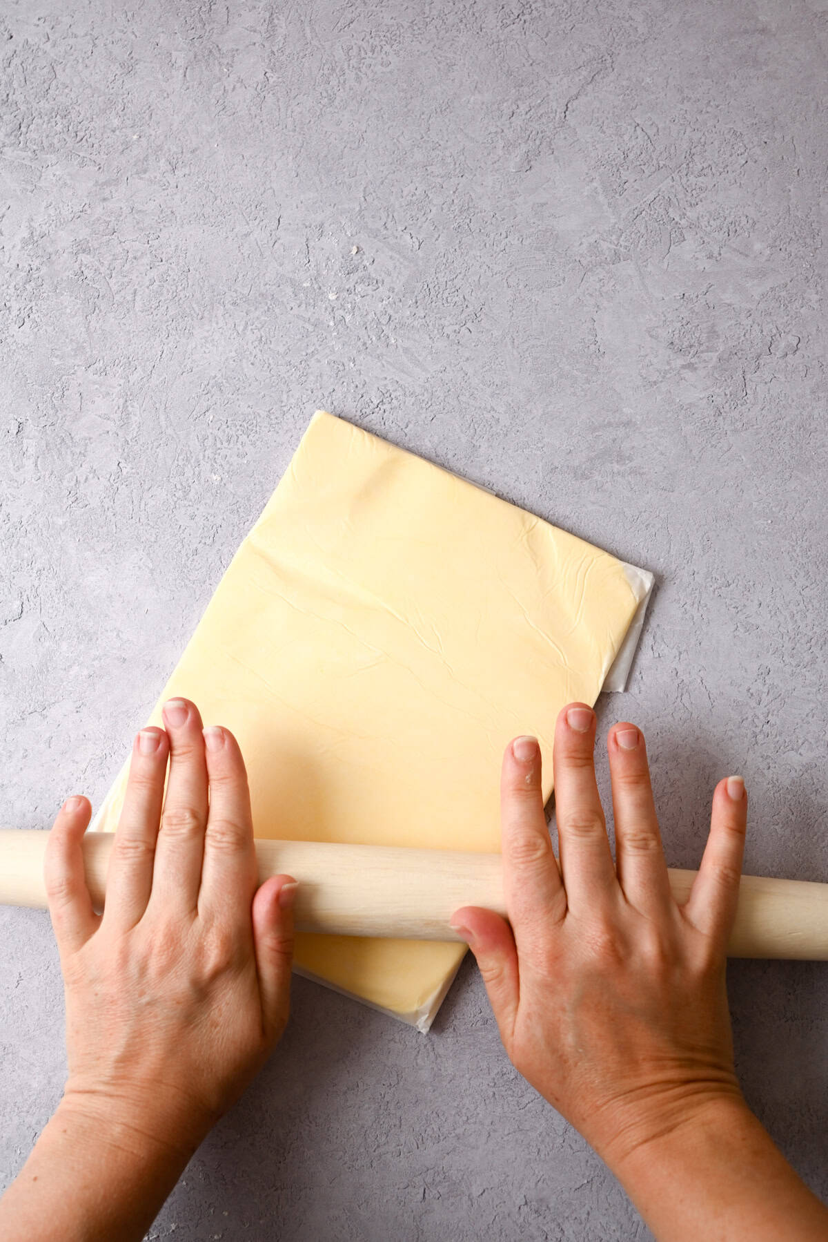 Two hands use a rolling pin to flatten pastry dough for sourdough pain au chocolat on a light gray textured surface.
