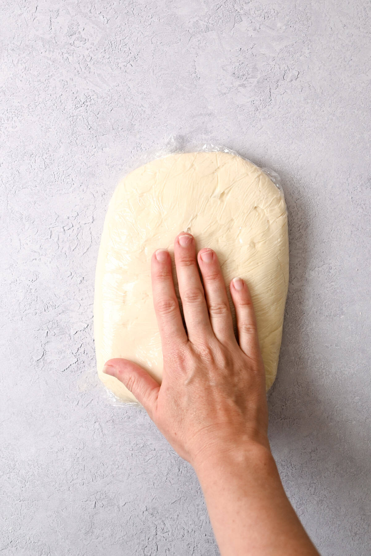 A hand presses down on a sheet of pale sourdough pain au chocolat dough wrapped in plastic wrap, resting on a light gray textured surface.