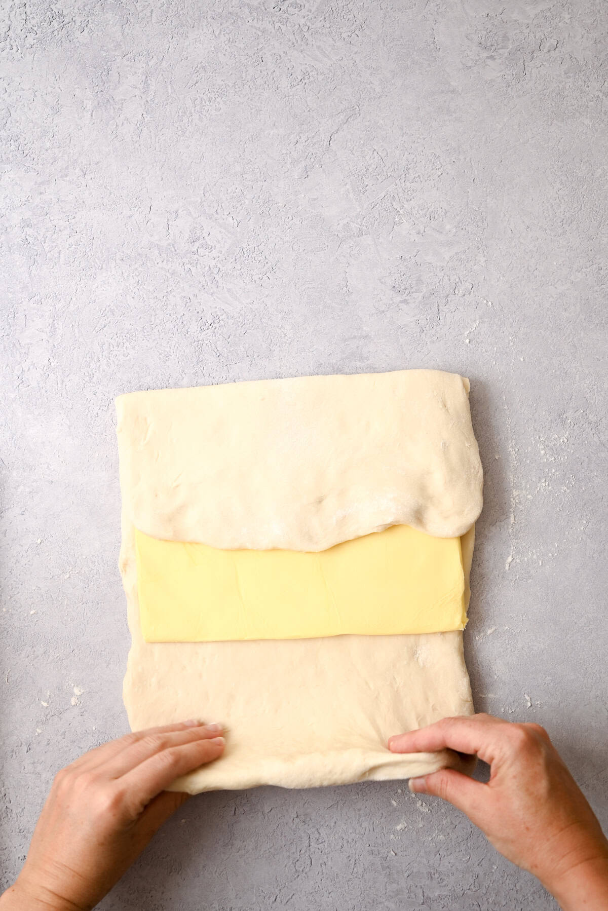 Hands folding dough over a rectangular slab of butter on a light gray, textured surface, preparing to laminate the dough for sourdough pain au chocolat.