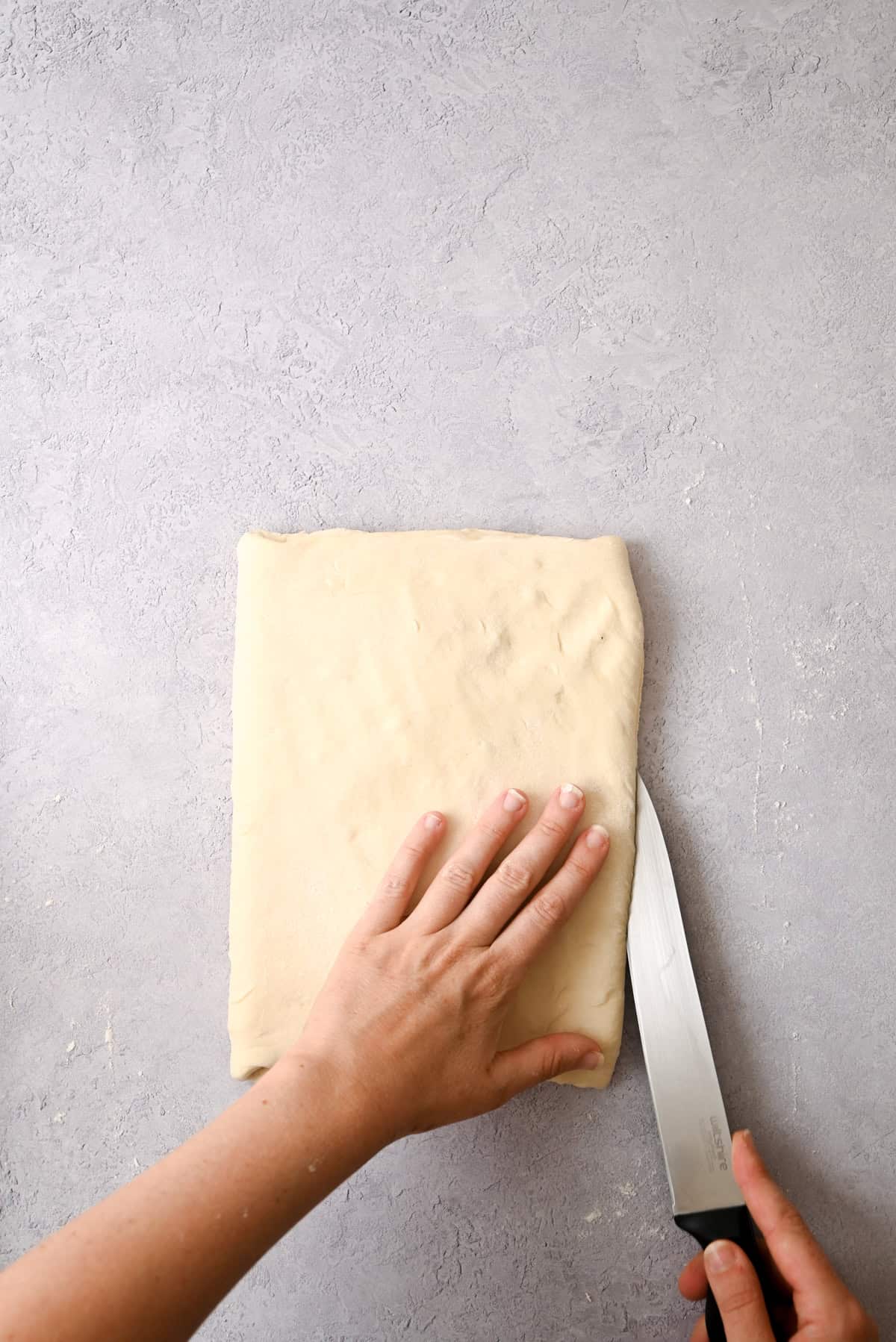 A person presses one hand on a folded sheet of sourdough pain au chocolat dough while slicing it with a large knife on a light gray surface.