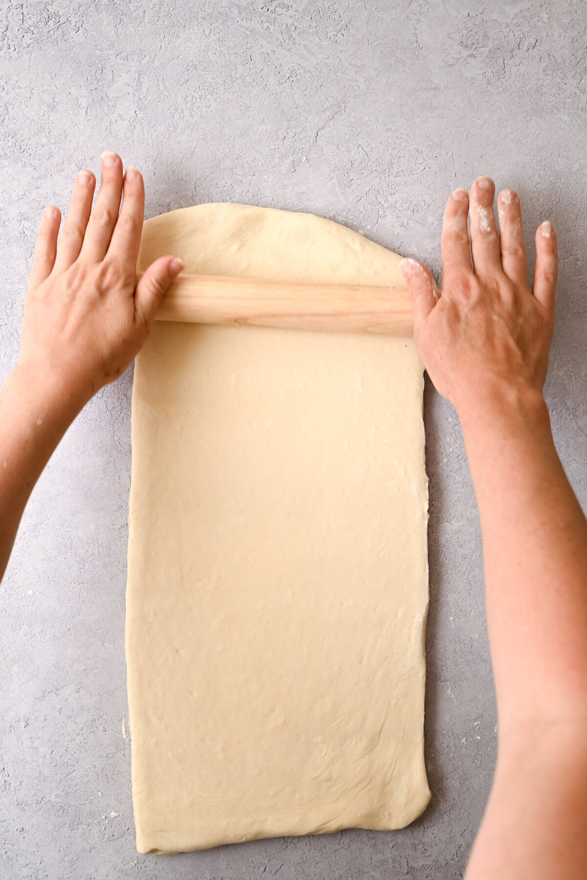 Two hands use a rolling pin to flatten a rectangular piece of sourdough pain au chocolat dough on a light gray surface.