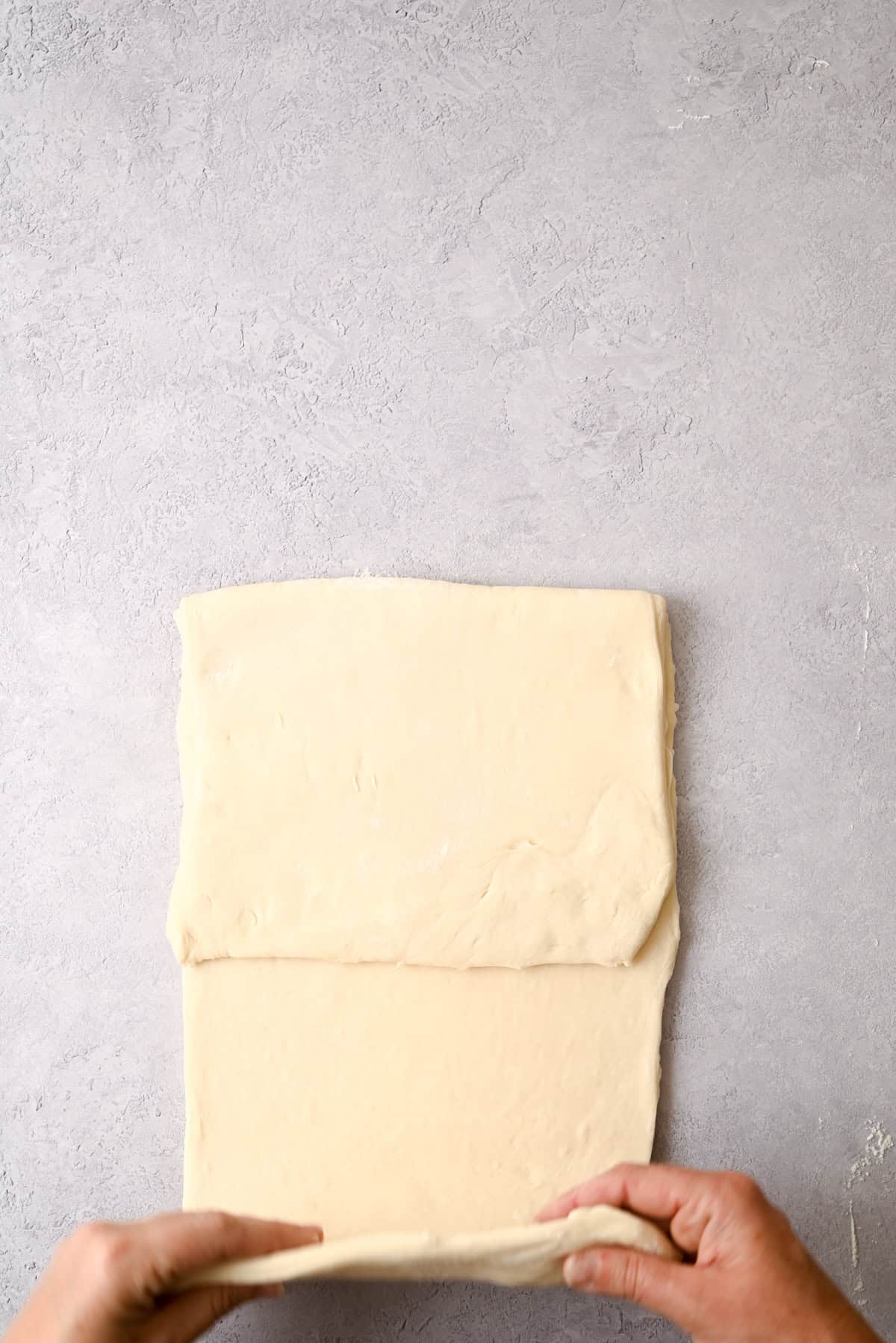 Hands are folding a rectangular piece of dough on a light gray surface, preparing it for baking a delicious sourdough pain au chocolat.