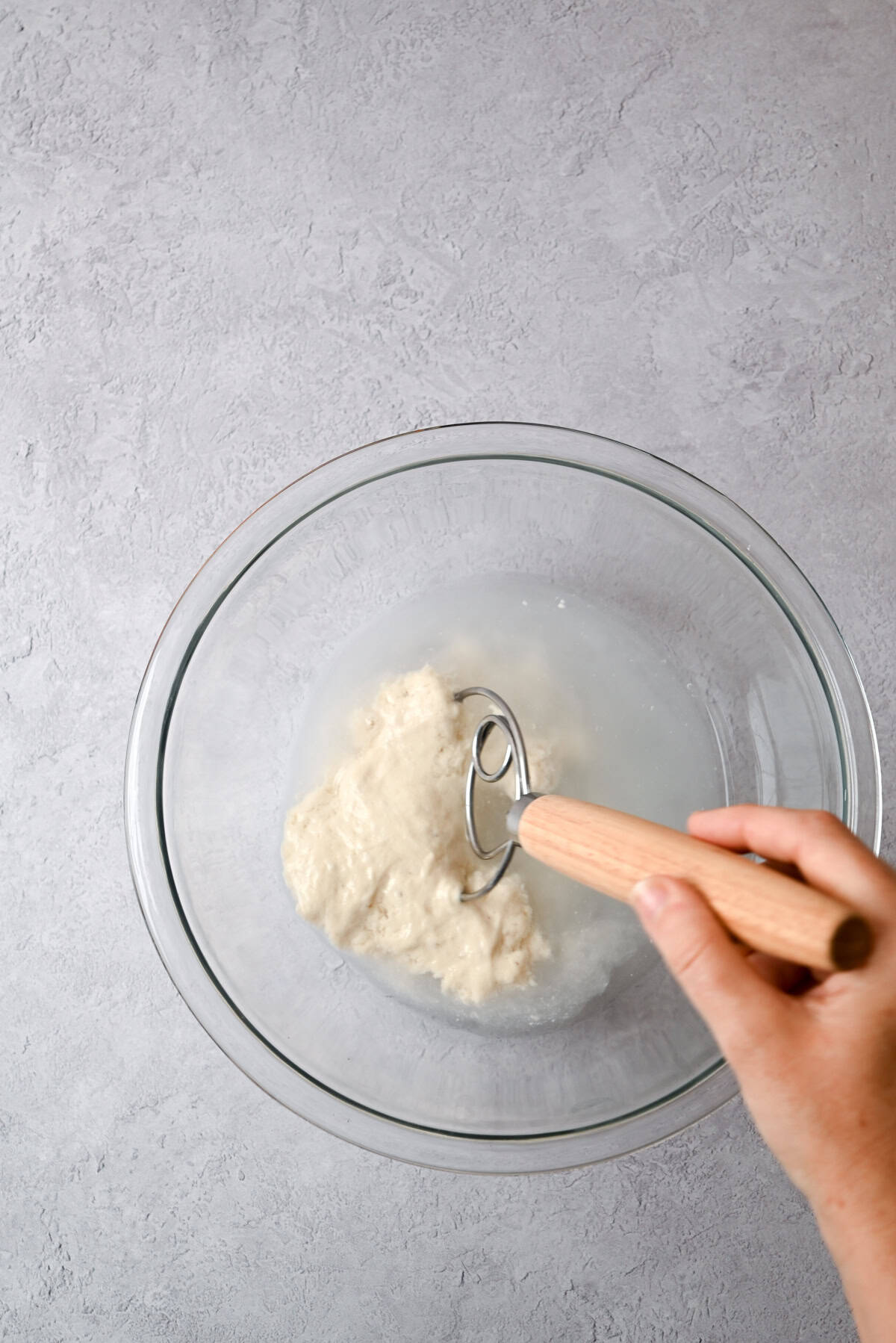 A hand holding a wooden-handled dough whisk is mixing dough and water for sourdough pain au chocolat in a clear glass bowl on a light gray surface.