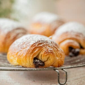 A close-up of a sourdough pain au chocolat dusted with powdered sugar, resting on a cooling rack with more croissants in the background. The setting appears light and airy with a soft focus.