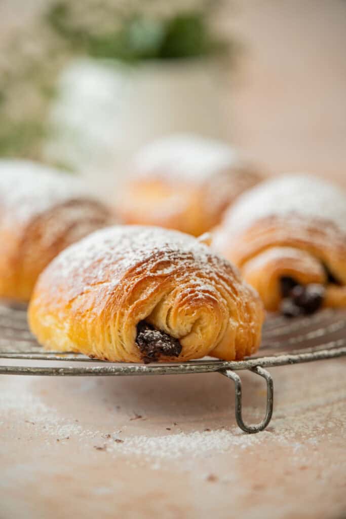 A close-up of a sourdough pain au chocolat dusted with powdered sugar, resting on a cooling rack with more croissants in the background. The setting appears light and airy with a soft focus.