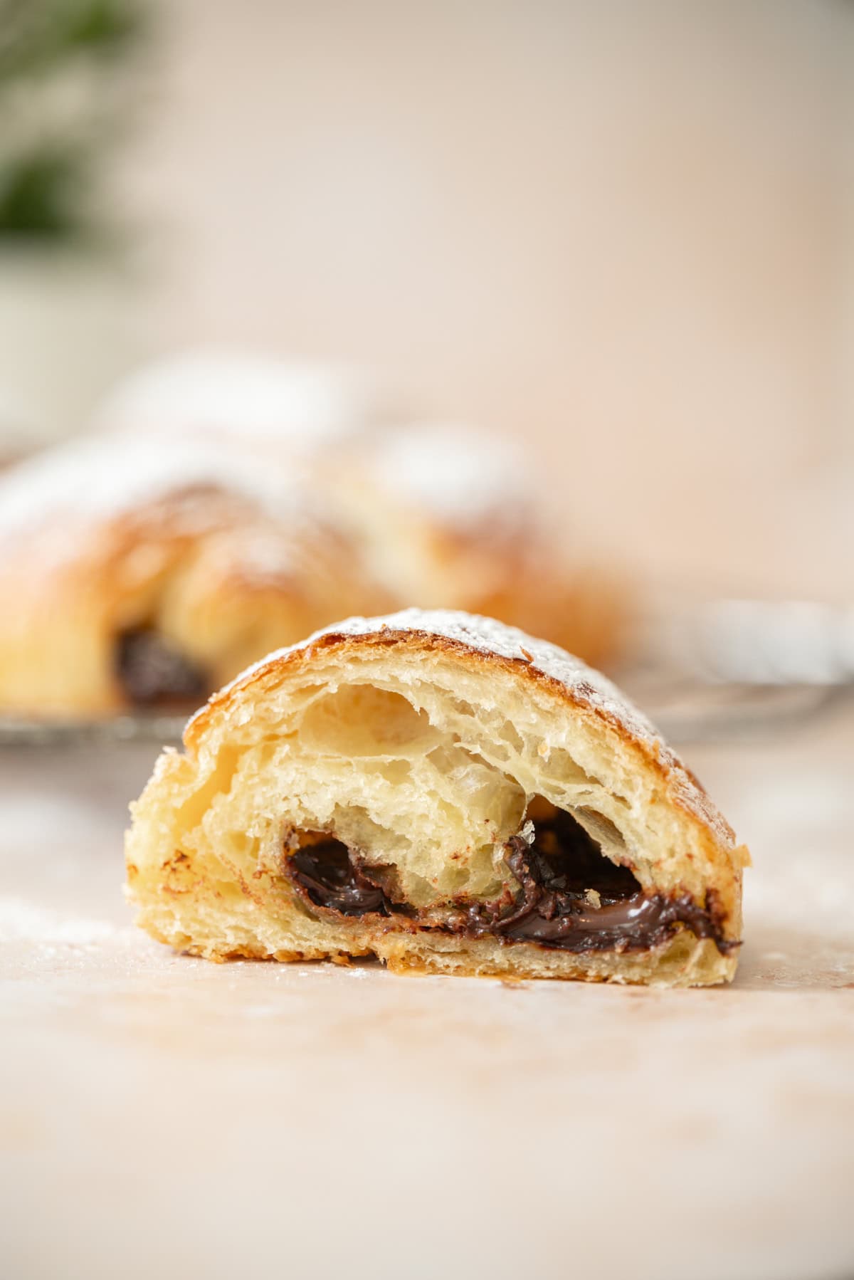 A close-up of a sliced sourdough pain au chocolat, showing its flaky, golden layers and melted chocolate filling, with a dusting of powdered sugar on top.