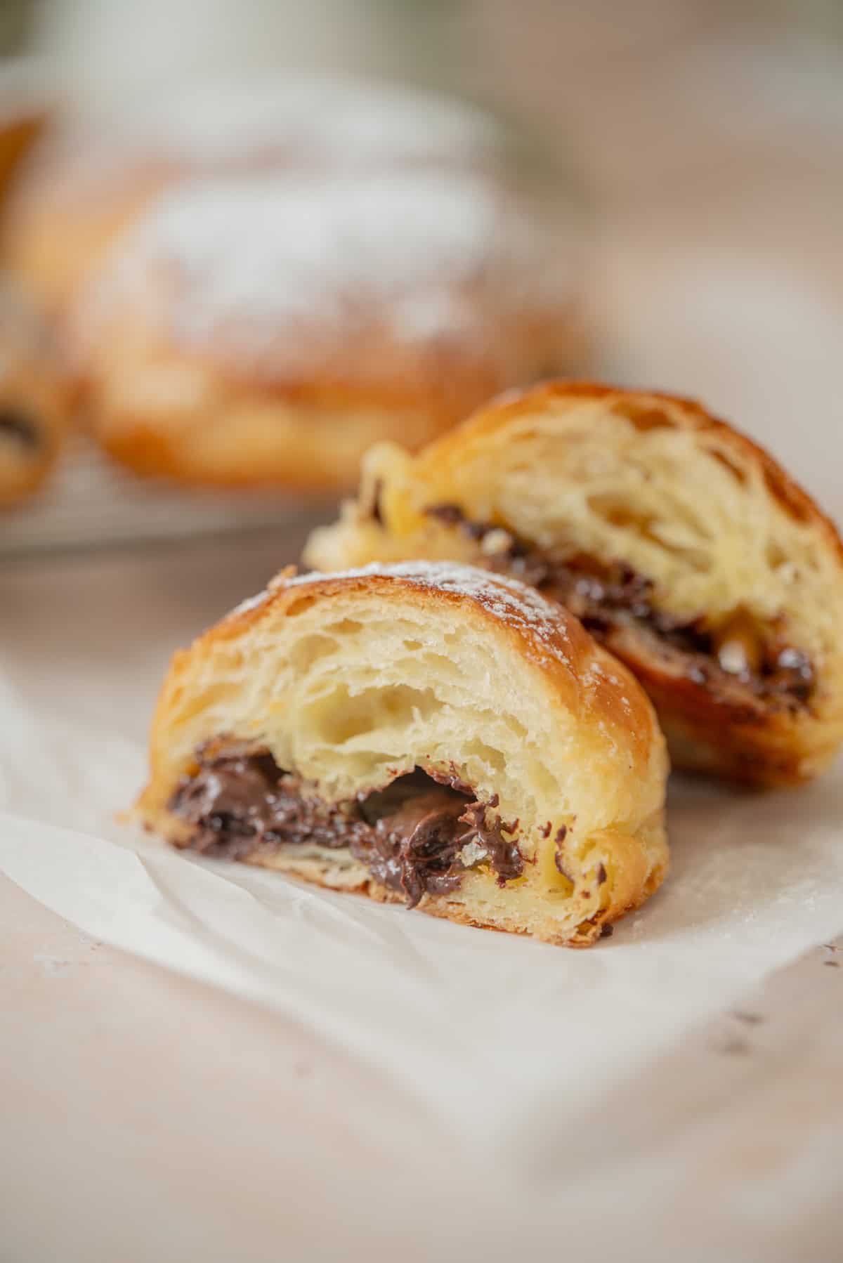 A close-up of a sourdough pain au chocolat cut in half, revealing its flaky layers and rich chocolate center, with powdered sugar dusted on top and another pastry blurred in the background.