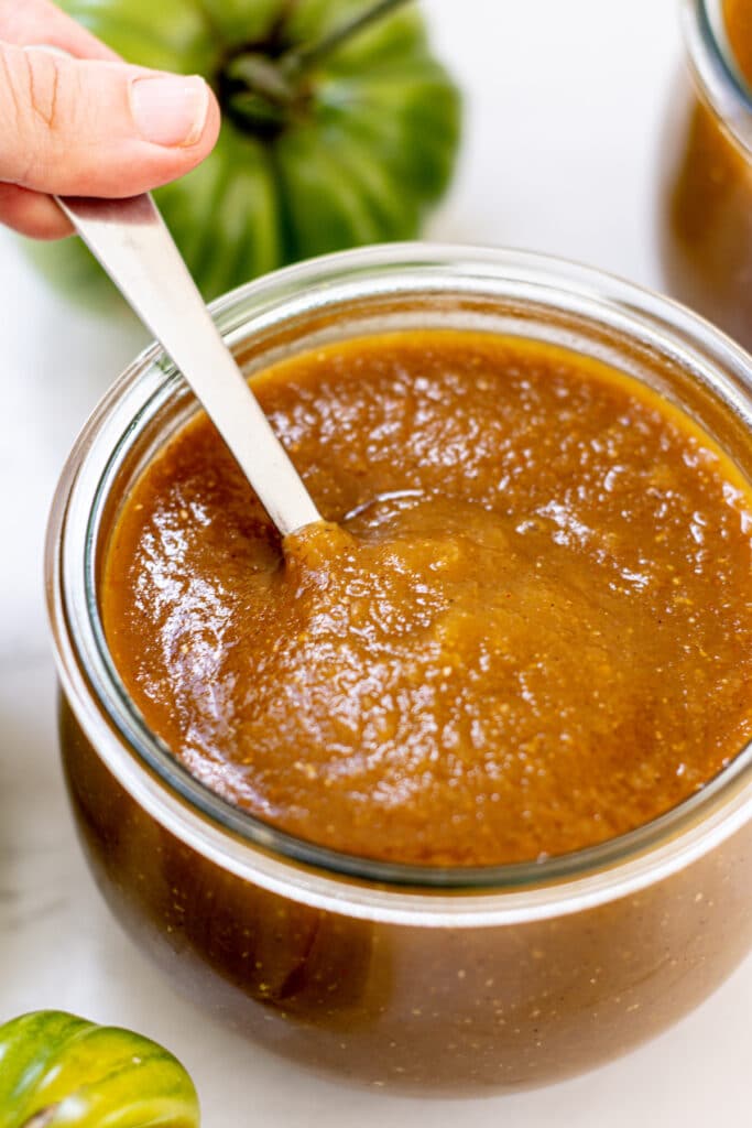 jar of barbecue sauce on a white bench surrounded by green tomatoes and a spoon being dipped into it