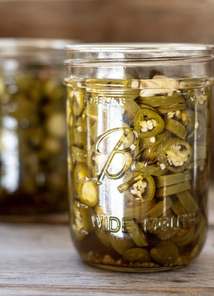 a close up of a glass jar of muted green jalapeño pepper slices on a wooden board.another jar is in the background