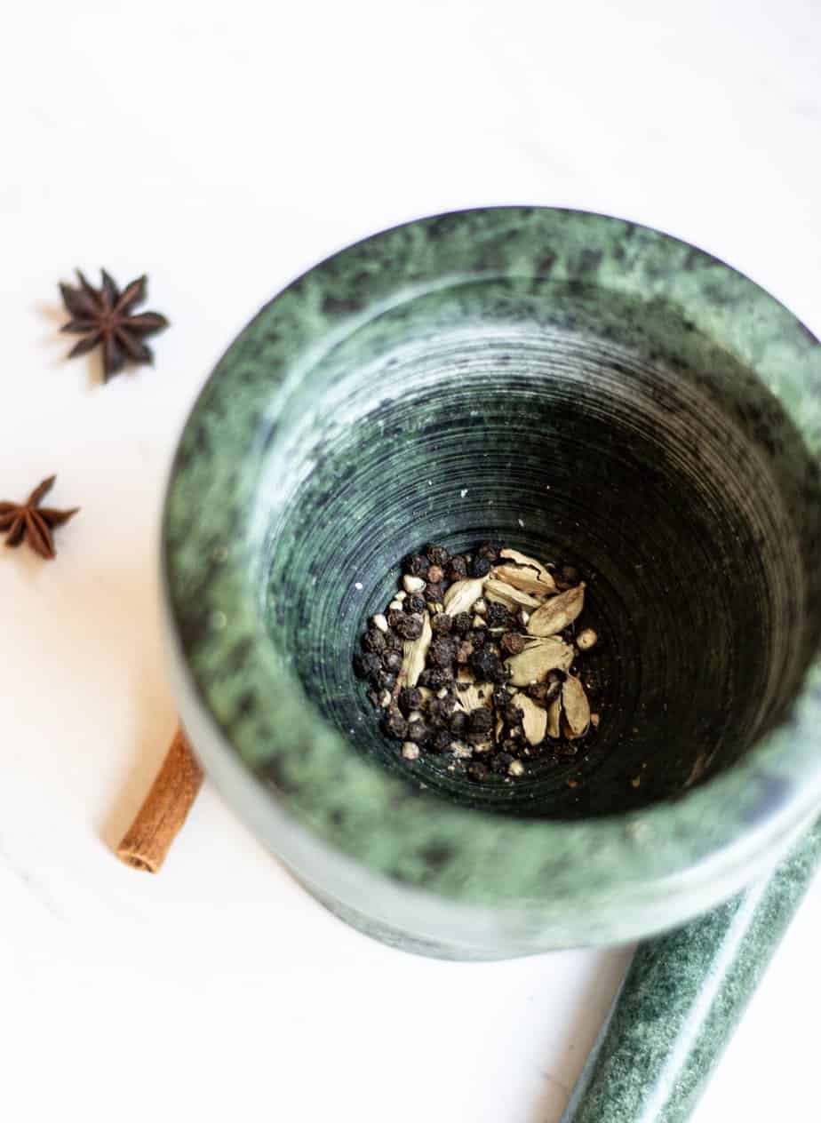 a green mortar with cardamom pods and peppercorns in it, a cinnamon stick and star anise in the background
