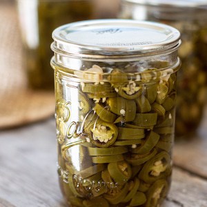 a glass jar of muted green jalapeño pepper slices on a wooden board. 2 other jars are in the background. Cropped picture