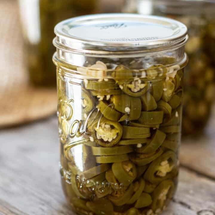 a glass jar of muted green jalapeño pepper slices on a wooden board. 2 other jars are in the background