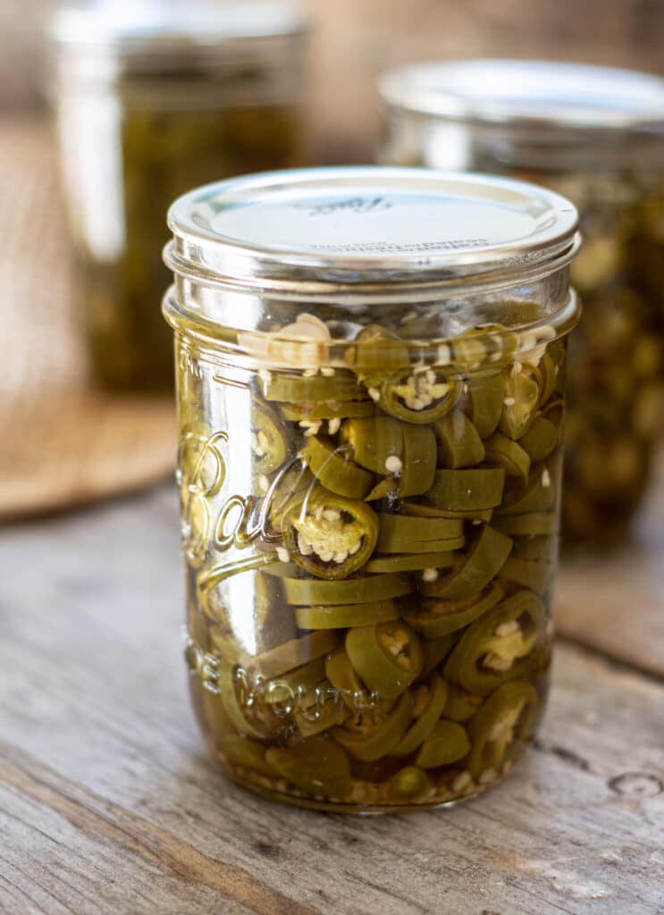 a glass jar of muted green jalapeño pepper slices on a wooden board. 2 other jars are in the background