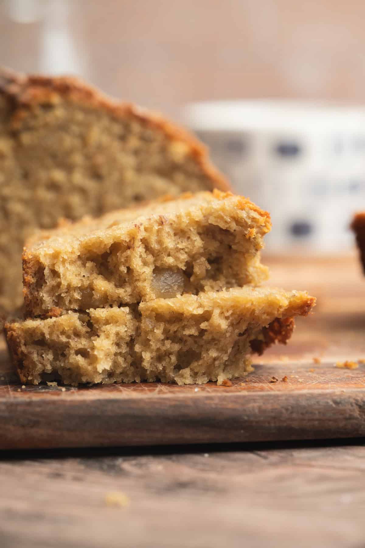 A close-up of two slices of moist Sourdough Banana Bread stacked on a wooden surface, with the main loaf and a blurred cup in the background.