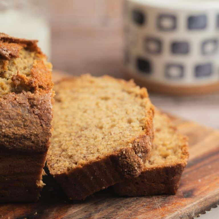 Close-up of sliced Sourdough Banana Bread on a wooden cutting board, with a blurred mug featuring a black-and-white geometric pattern in the background.