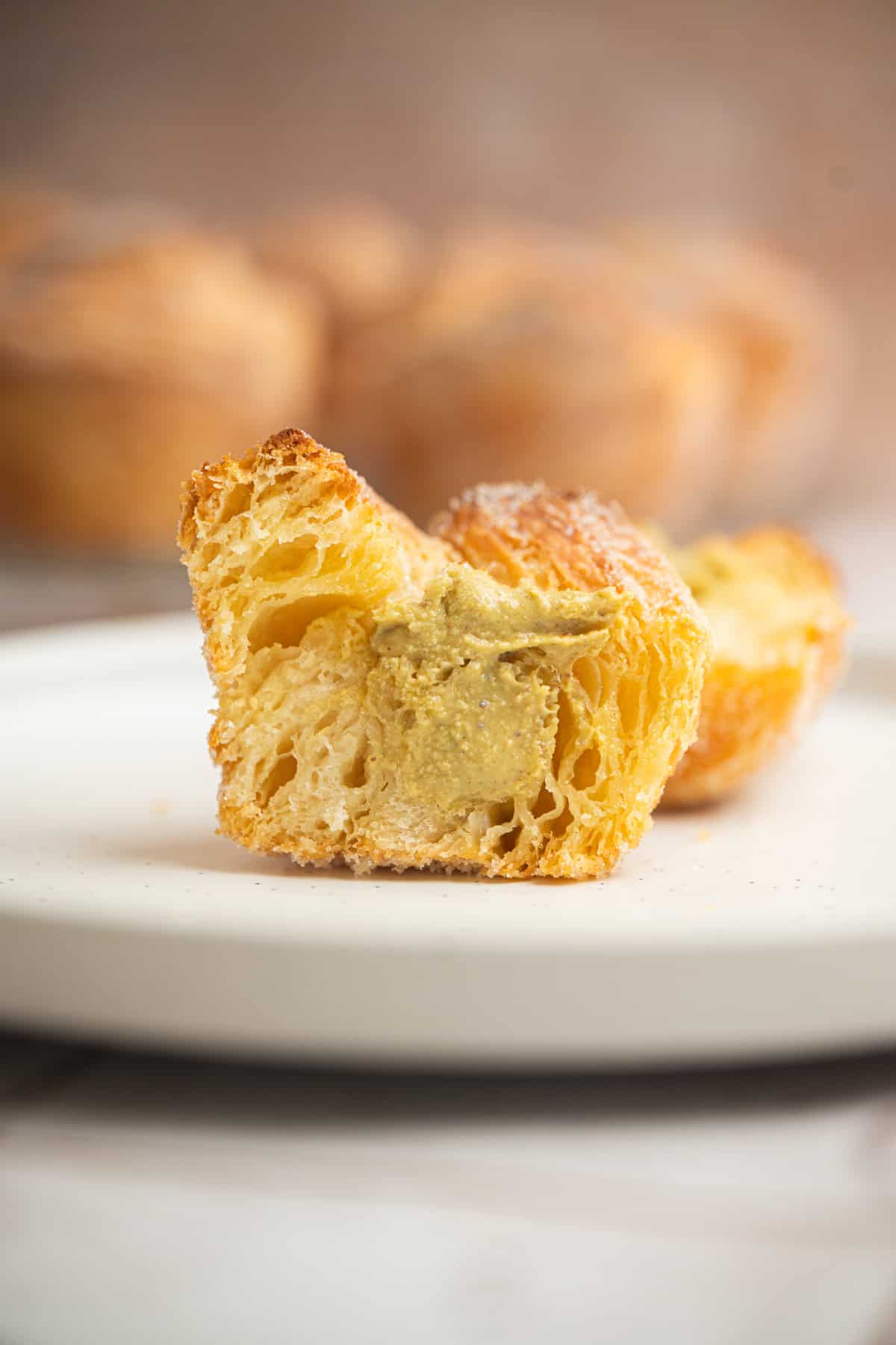 A close-up of a flaky, golden-brown sourdough cruffin with creamy filling visible inside, placed on a white plate. The cruffin’s airy, layered texture is perfectly highlighted.