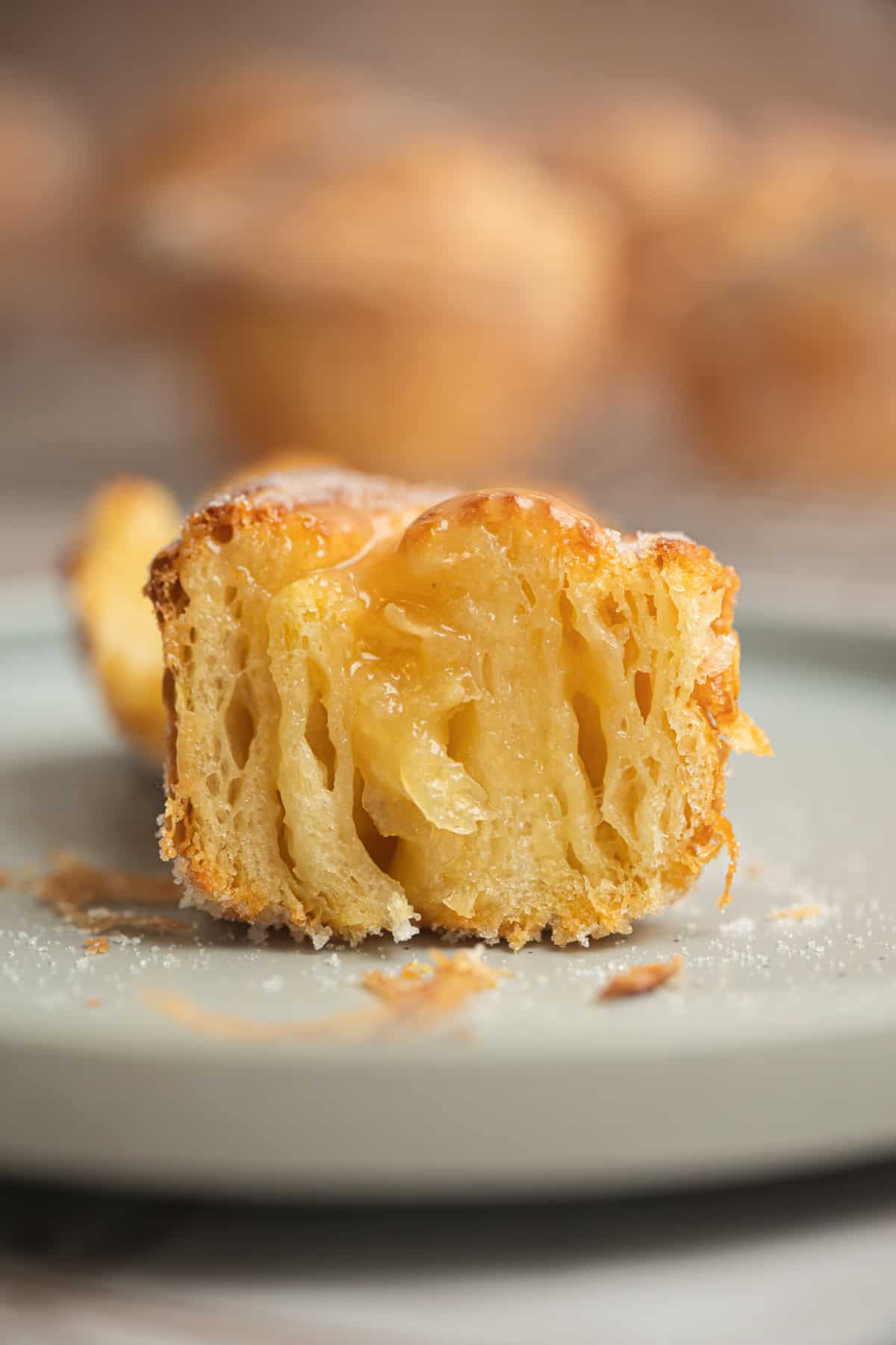 A close-up of sliced sourdough cruffins reveals a golden, flaky pastry with a caramelized, layered interior on a light gray plate, crumbs scattered around. The softly blurred background adds warmth.