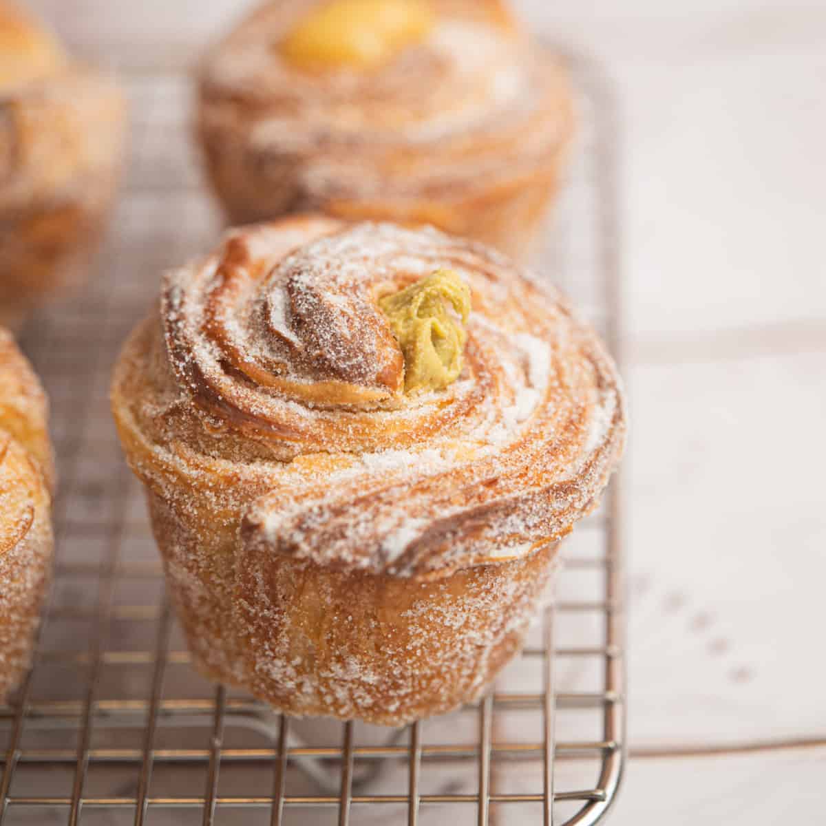 A close-up of a sugar-dusted, swirled sourdough cruffin with a creamy filling on top, resting on a wire cooling rack. Other similar pastries are slightly out of focus in the background.