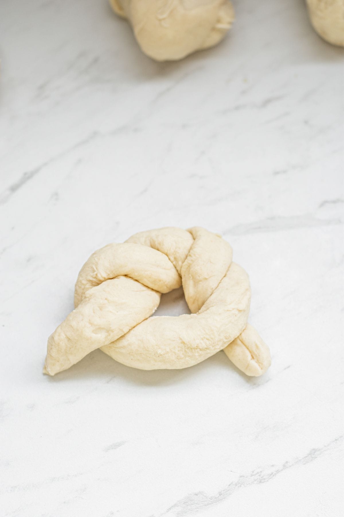A piece of uncooked sourdough garlic knot dough shaped into a knot rests on a white marble surface, with more dough pieces partially visible in the background.