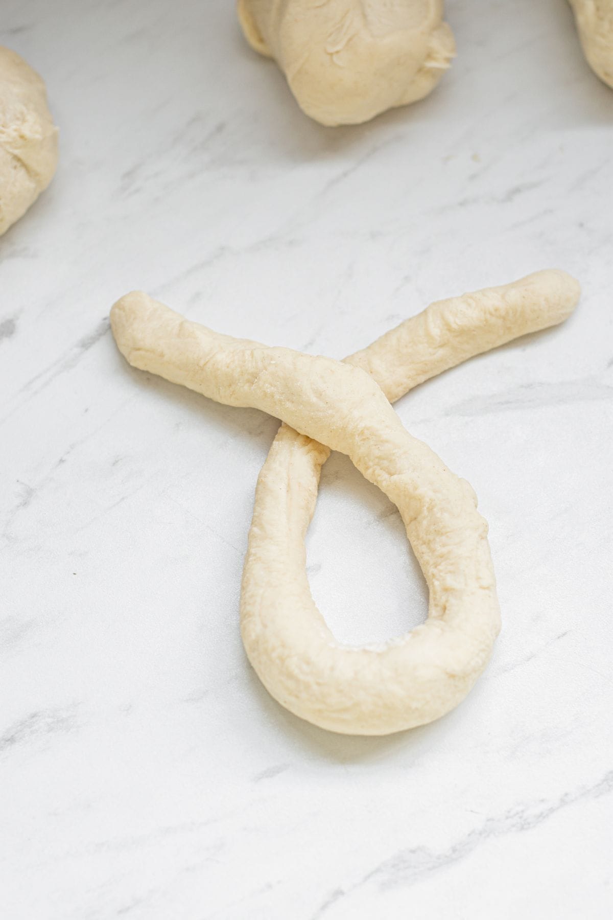 Unbaked pretzel dough shaped into a loop with crossed ends, resting on a white marble surface alongside sourdough garlic knots and additional dough pieces in the background.