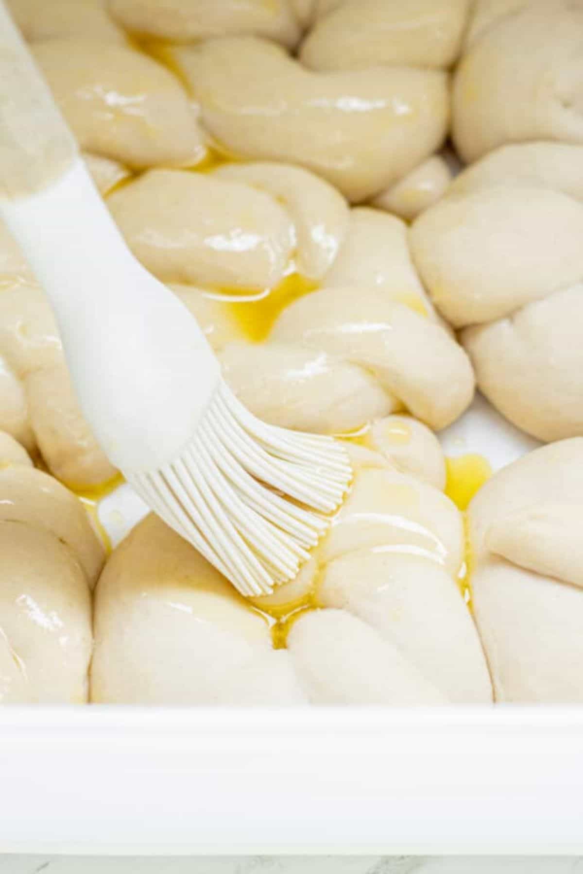 A pastry brush is spreading egg wash over unbaked sourdough garlic knots, braided and arranged neatly in a white tray.