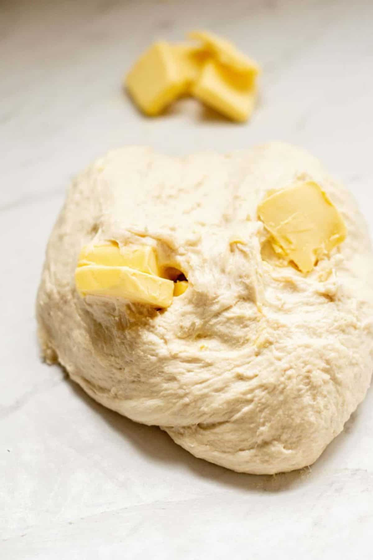 A mound of raw dough for sourdough garlic knots with two chunks of butter pressed into it sits on a light-colored surface, with extra butter pieces in the background.