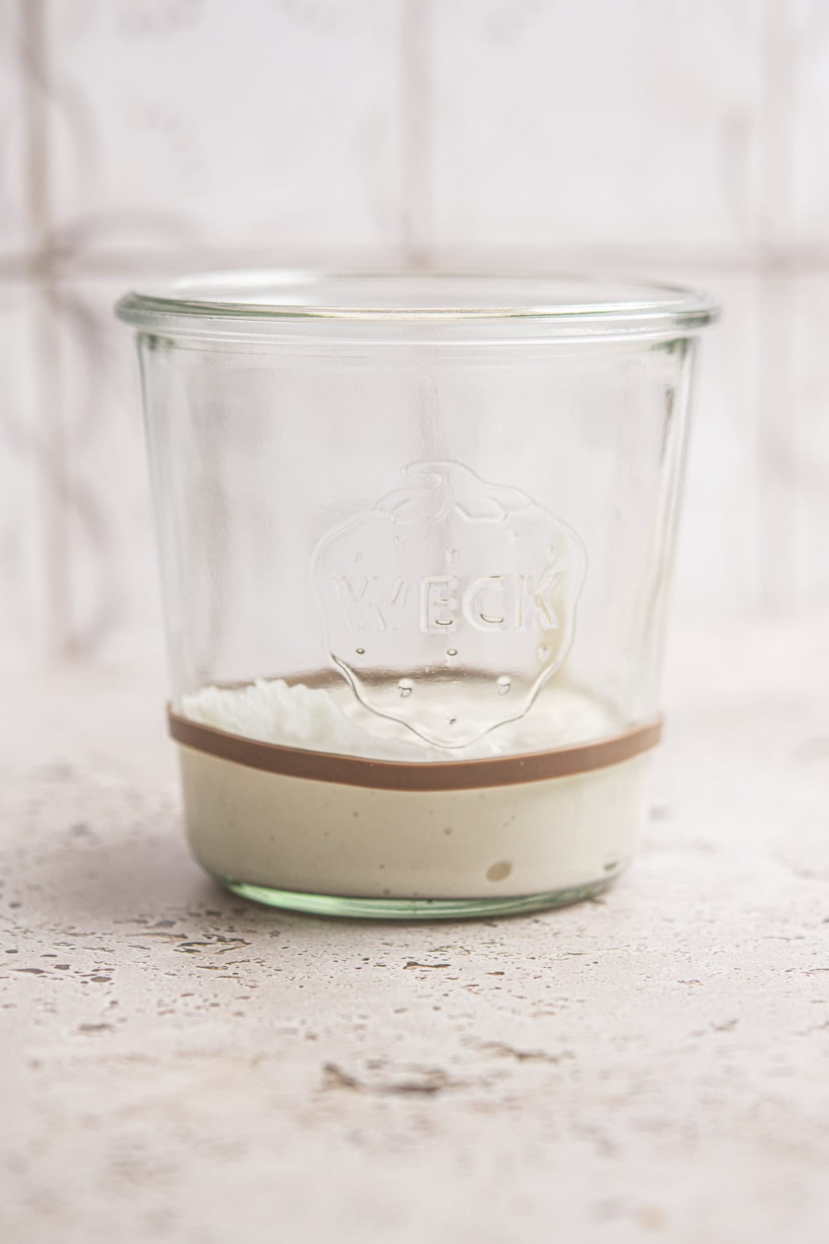 A glass jar with a small amount of creamy, beige sourdough starter resting at the bottom sits on a light textured surface, hinting at careful sourdough starter maintenance. The jar is labeled WECK and the background is softly blurred.