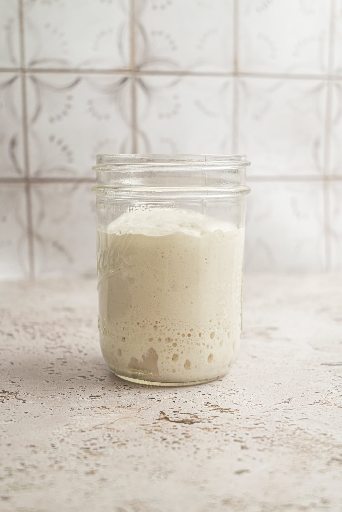 A glass jar filled with bubbly, creamy sourdough starter sits on a light textured surface with a patterned tile background, showcasing the results of careful sourdough starter maintenance.