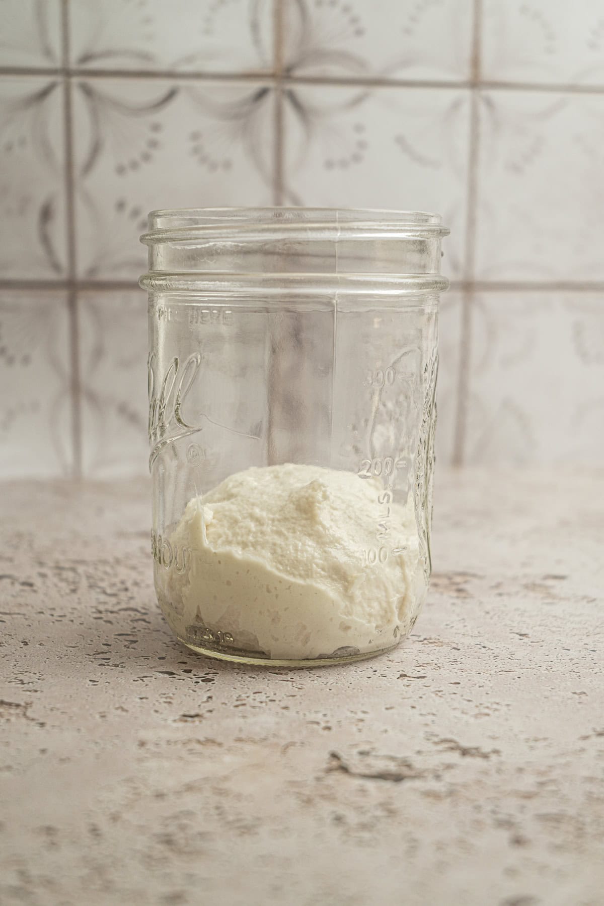 A glass jar containing a small amount of dough—perfect for sourdough starter maintenance—sits on a light, textured surface with a patterned tile background.