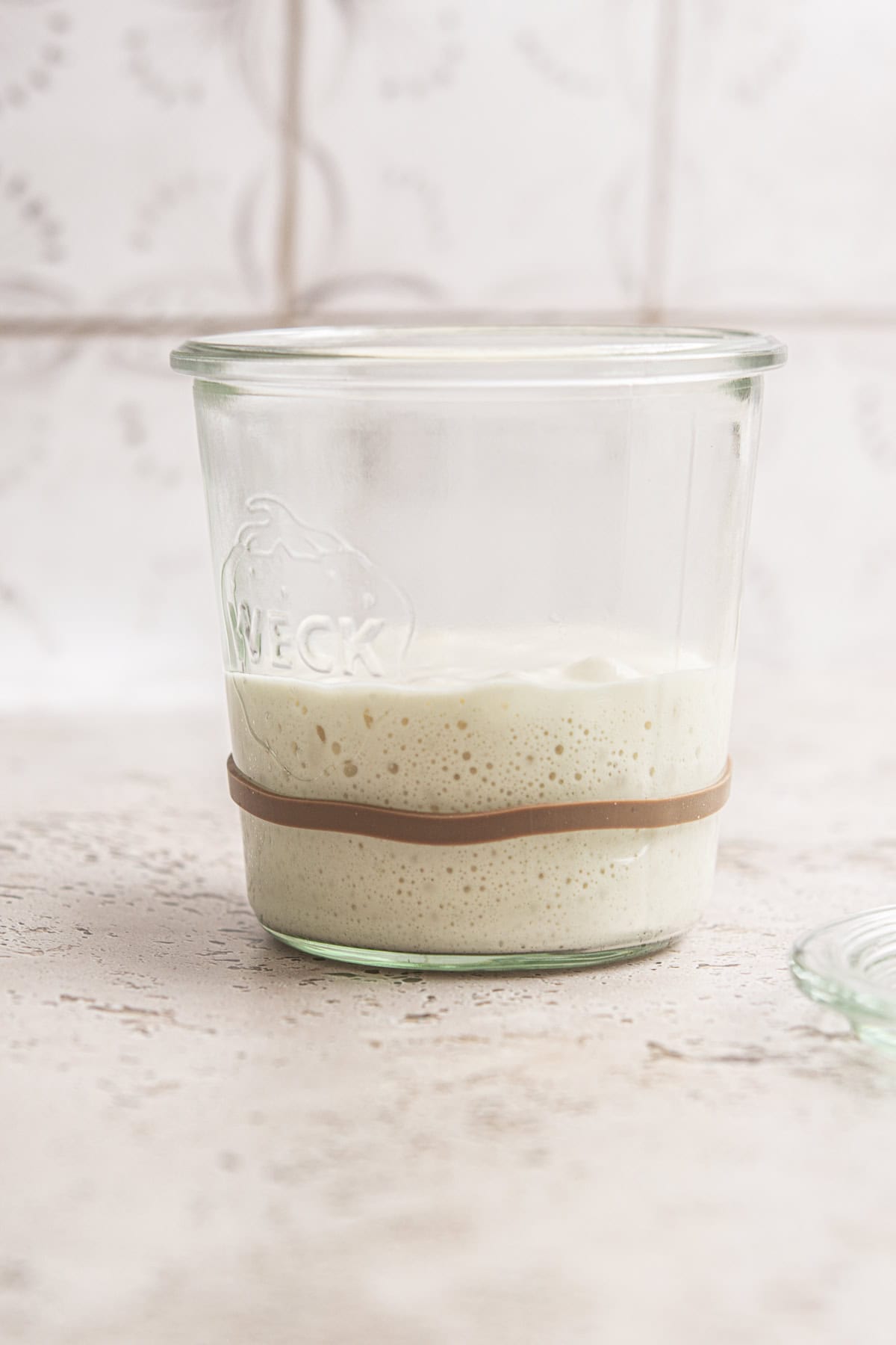 A glass jar with sourdough starter sits on a light textured surface, marked with a rubber band to track sourdough starter maintenance. The bubbly mixture rises inside, while the background features a softly blurred tile.