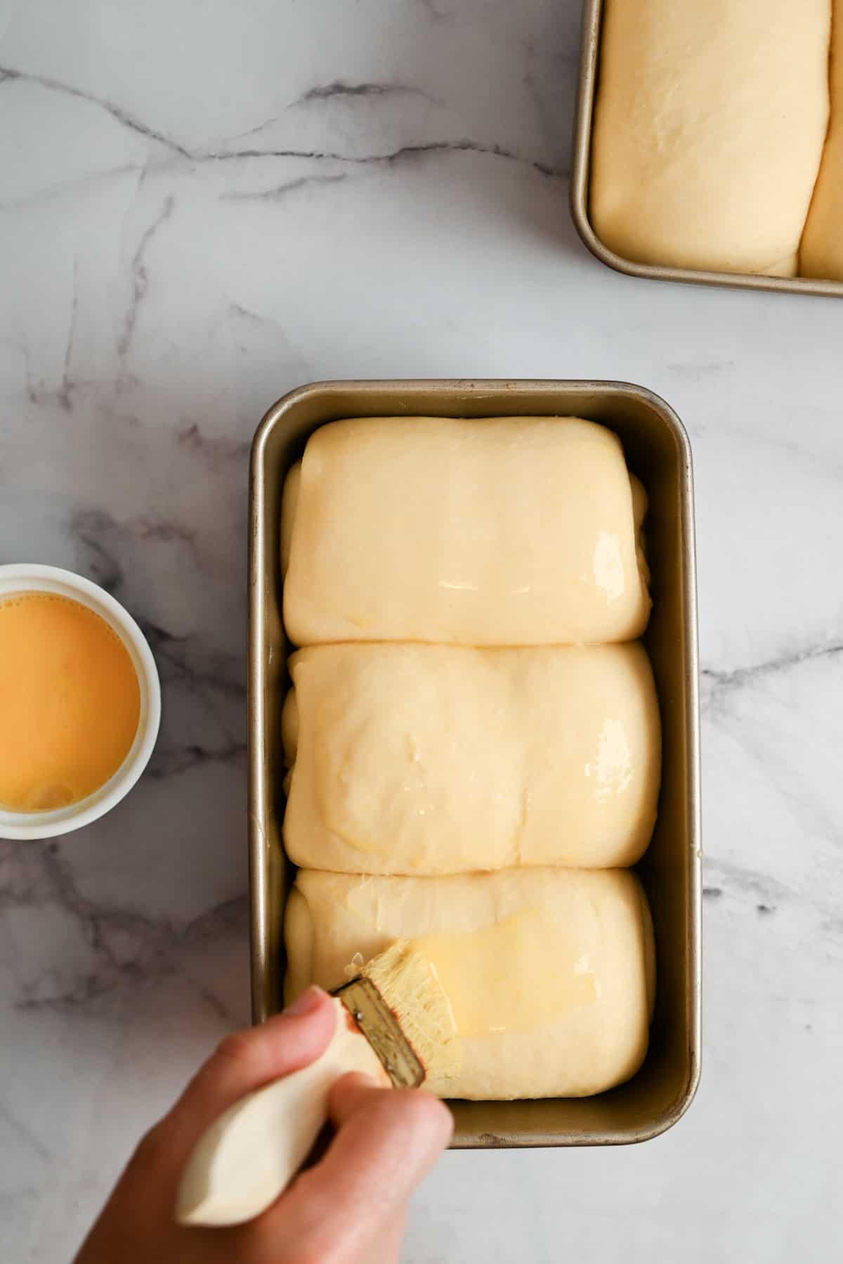 A hand brushes egg wash onto three unbaked sourdough milk bread rolls in a loaf pan. A small bowl of egg wash and another loaf pan with dough rest on a marble surface.