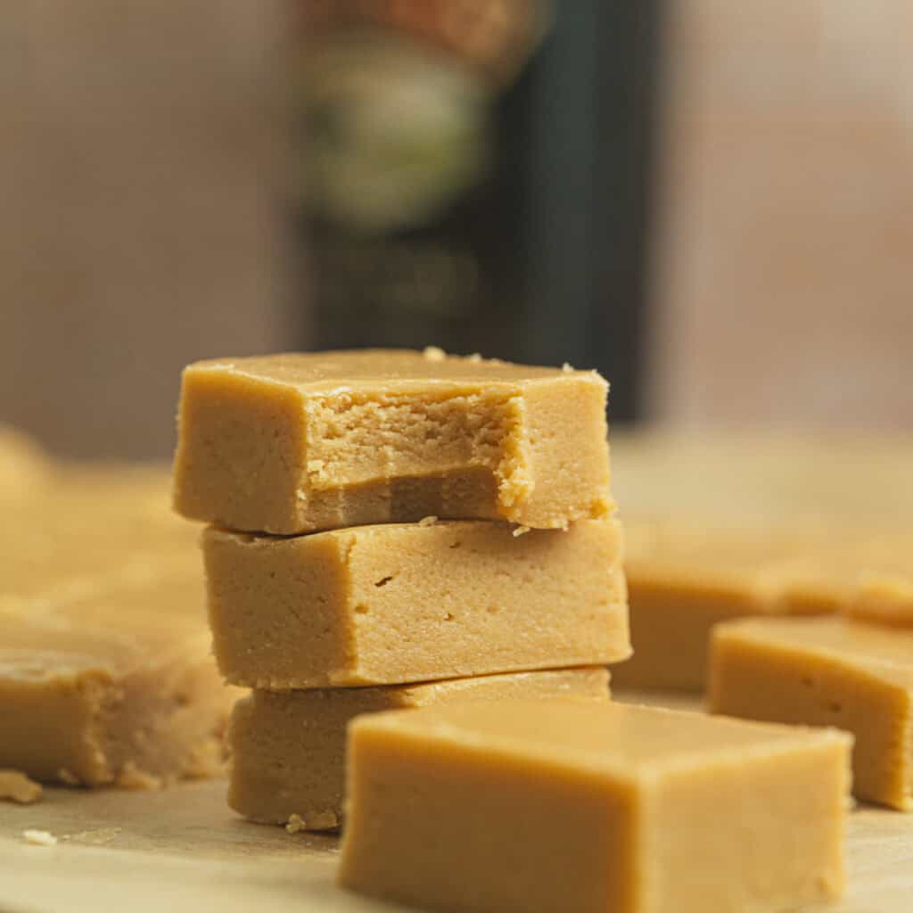 Three pieces of creamy, light brown Baileys Fudge are stacked on top of each other, with the top piece showing a bite taken out. More pieces of fudge are visible in the blurred background.