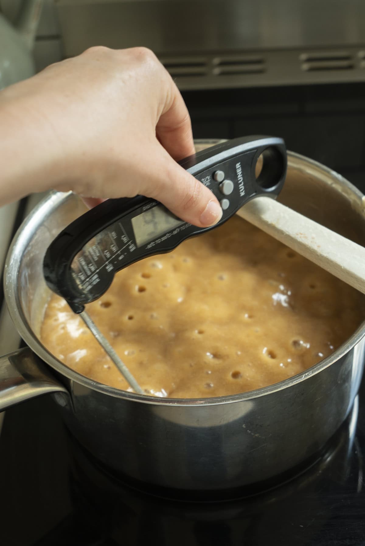 A hand holds a digital thermometer over a bubbling, caramel-colored Espresso Fudge mixture in a stainless steel pot on the stove, with a wooden spoon resting on the pot’s edge.