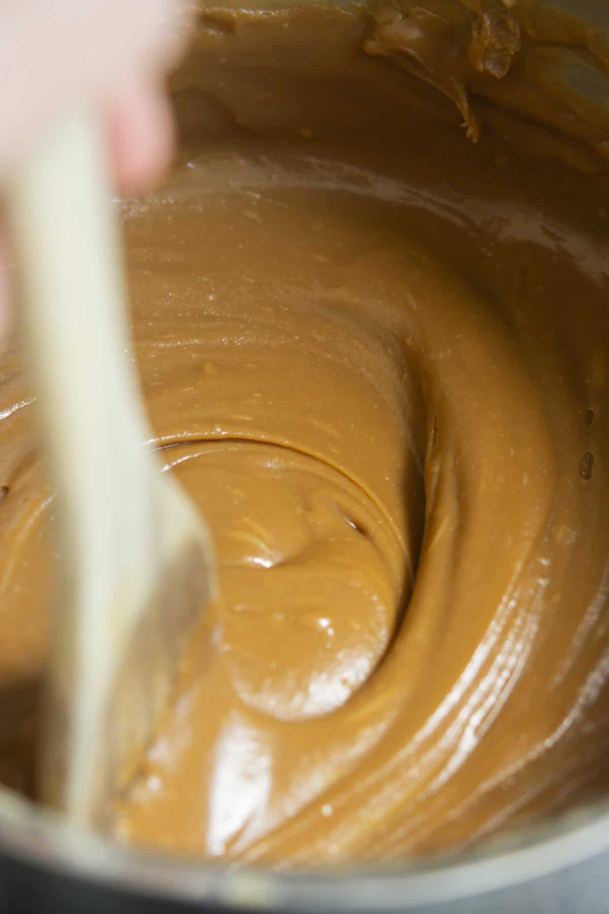 A close-up of a thick, creamy Espresso Fudge caramel mixture being stirred with a wooden spatula inside a metal pot.
