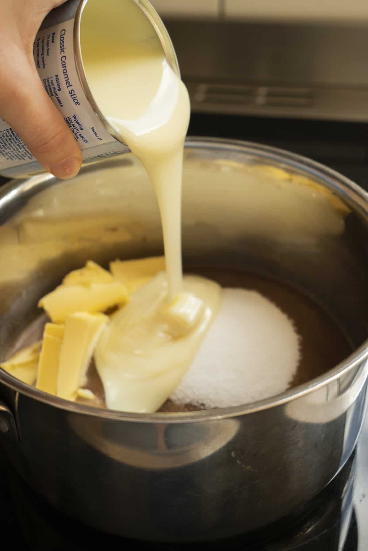 A hand pours sweetened condensed milk from a can into a saucepan with butter and granulated sugar, preparing ingredients for rich Espresso Fudge.