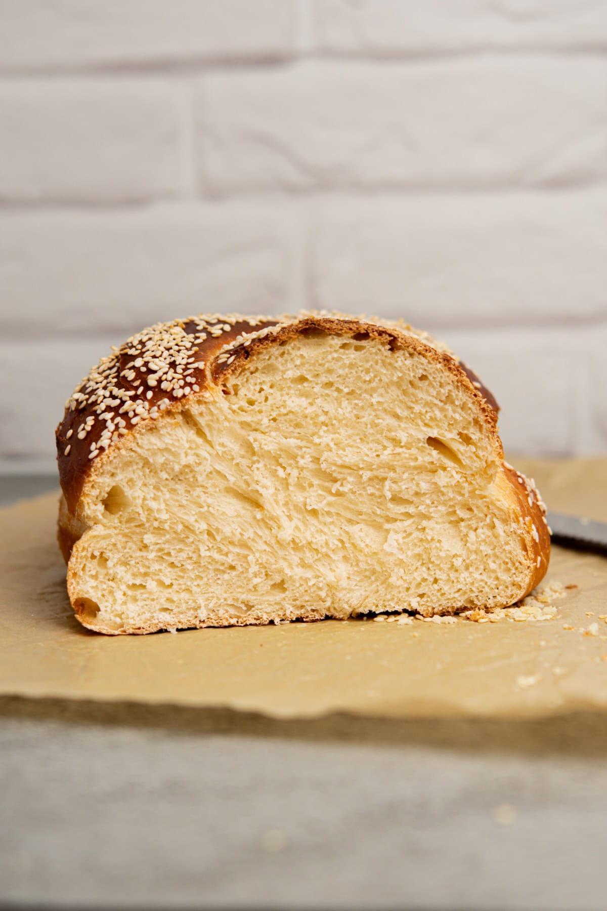 A sliced loaf of sourdough challah bread with a golden crust and sesame seeds on top, resting on a piece of parchment paper. The background features a white brick wall.