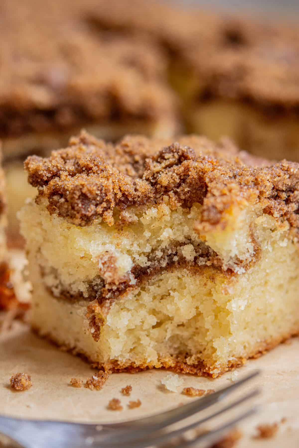 A close-up of a slice of sourdough coffee cake with a crumbly cinnamon topping and visible swirl. The cake is moist and fluffy, with a fork shown in the foreground and more cake pieces blurred in the background.